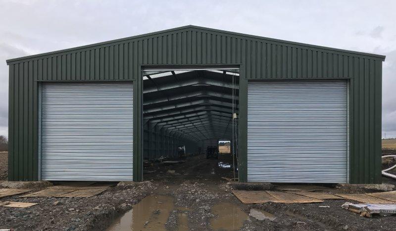 A symmetrical front view of a large olive green steel warehouse with two closed silver roller doors and one open central bay showing the interior frame.